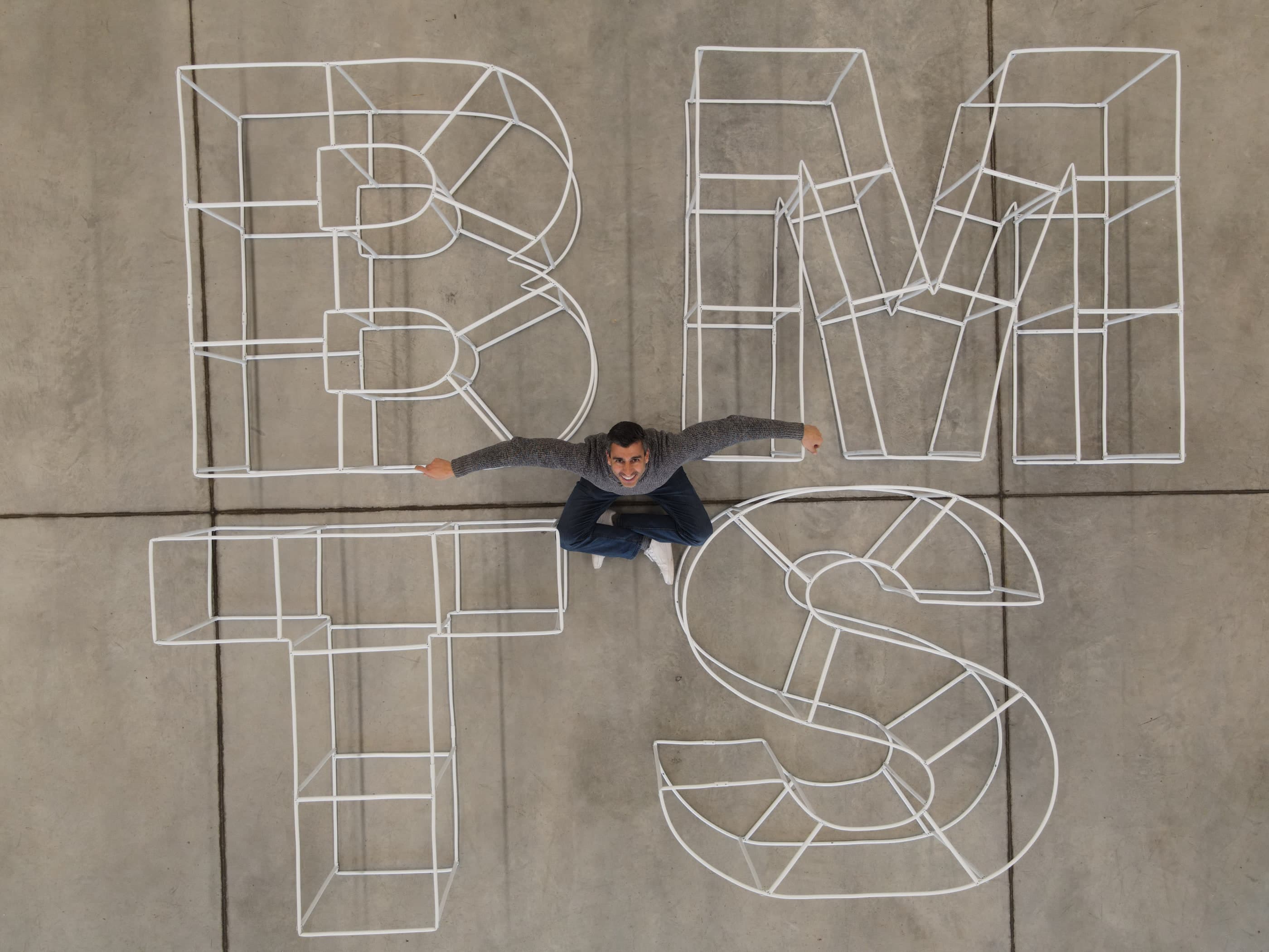 Vista aérea de varias letras gigantes de tubo blanco sobre suelo de hormigón, con dos personas sentadas dentro de ellas que dan la escala.
